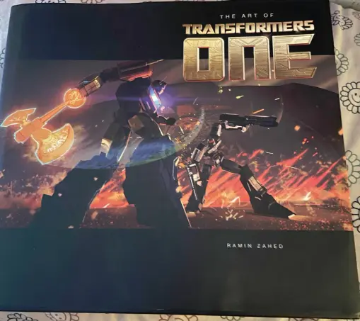 The book is a big square artbook called "The Art of Transformers One". It has a picture of Optimus Prime and Megatron fighting in a battle surrounded by fire on the cover.