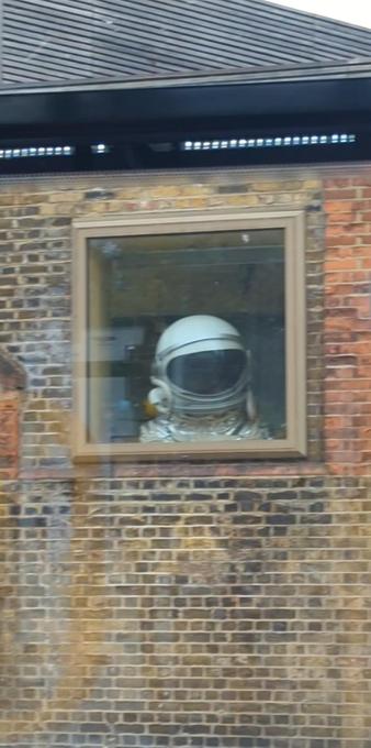 A "London yellow brick" wall with window positioned close to roofline. An astronaut's closed helmet and upper part of suit positioned to look like someone is looking out and to the right of the window.