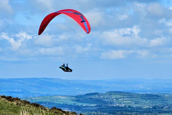 A paraglider with a red and blue canopy soaring over a green landscape with rolling hills and a cloudy sky.