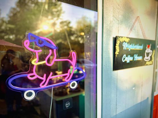 A pink, purple, and yellow neon sign of a dachshund dog smiling and wearing sunglasses while on a skateboard displays in a window next to an exterior wall with a wooden sign hanging that reads, "Neighborhood Coffee House."