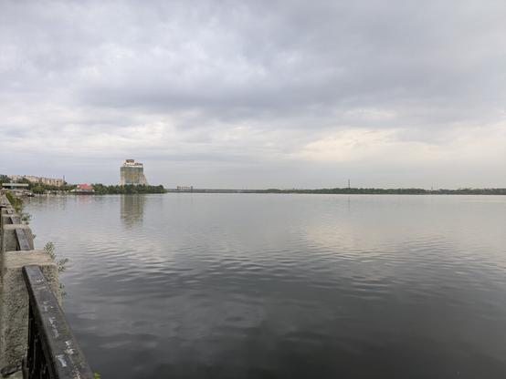 The image shows a view across a wide body of water towards the skyline of Dnipro, Ukraine. In the center of the image, prominently featured in the distance, is the unfinished Hotel Parus, a tall, white building with a distinctive shape. The hotel rises above the other buildings visible on the shore. The water in the foreground is calm, reflecting the overcast sky. On the left side of the image, a concrete embankment with a dark railing is visible, suggesting the photo was taken from the waterfront. The overall impression is one of a somewhat grey day, with the imposing structure of the Hotel Parus standing out against the cloudy sky.