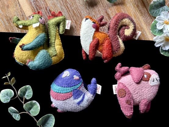 Four handmade felt plushies photographed on a black and wooden background with a few decorative plants around it. There is a crocodile, a squirrel, a pigeon, and a pig.