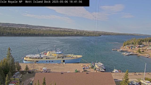 The Mott Island dock and the passenger vessel that ferries park visitors in Isle Royale National Park fill the foreground of this image, with water and a tree-covered landscape under a blue sky as a backdrop.