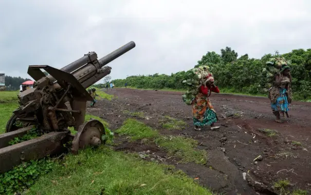 Two women carry heavy loads of freshly picked cabbages next to a rusting artillery piece.
