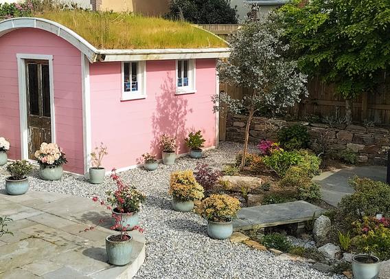 Photograph of a back garden.

The main feature, slightly left of centre, is a bright pink shed with a barrel-shaped green roof. There are two windows down the side, and an old door from which the paint has peeled.

On the left of the picture is a circular sandstone patio, and on the right is a flat slate bridge over a dry stream bed. Along the back is a stone wall.

There are numerous grey ceramic pots planted with a variety of small shrubs, and larger shrubs and trees dotted around. The ground is laid with grey gravel.