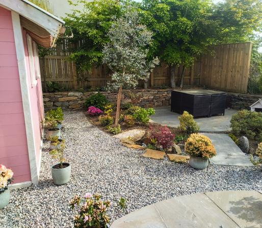 The same garden as in the previous picture, but taken from a different angle and pointed at a different corner.

The edge of the pink shed is visible in the left of the photo, and the flat slate bridge is seen to lead to another circular patio, this one laid with slate, on which a square table and chairs set is sitting.