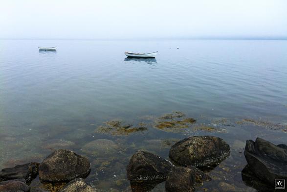 Color photo looking out from a rocky shoreline across calm waters with two small boats moored offshore. In the distance is a fog-shrouded hazy shoreline.