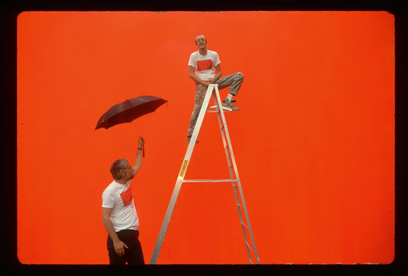 Keith Haring on a ladder, in front of the bright orange undercoat on which he would paint his mural Crack is Wack, with the New York Park Commissioner Henry Stern holding an umbrella