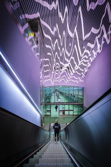 A girl is going down an elevator under zigzagging pink neon lights towards a platform bathed in blueish tones at Odenplan metro station in Stockholm. Her dark silhouette is slightly blurry due to a long exposure.