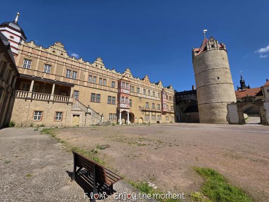 Das Bild zeigt den Innenhof von Schloss Bernburg in Sachsen-Anhalt an einem sonnigen Tag mit klarem, blauen Himmel. Das Schlossgebäude nimmt den größten Teil des Hintergrunds ein. Es handelt sich um einen langgestreckten Renaissancebau aus hellem Sandstein mit zahlreichen, gleichmäßig angeordneten Fenstern. Die Fassade ist reich gegliedert mit Giebeln und roten Zierleisten.

Links führt eine breite Steintreppe mit einem Geländer zu einer erhöhten Galerie mit Rundbögen. Rechts neben dem Hauptgebäude steht ein großer, runder Turm mit schlichter Fassade. Dieser Turm ist als Eulenspiegelturm bekannt und ein historischer Teil des Schlosses. Er trägt eine goldene Wetterfahne auf dem Dach.

Der Boden des Innenhofs ist teils gepflastert, teils mit festem Erdreich bedeckt, aus dem vereinzelt Gras wächst. Im Vordergrund steht eine einfache, hölzerne Parkbank. Am unteren Rand des Bildes ist der Schriftzug „F.loki – Enjoy the moment“ zu lesen.