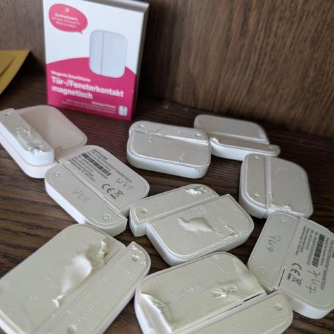 Mehrere weiße Tür-/Fensterkontakte mit Kleberesten liegen auf einer Holzfläche neben der Produktverpackung.