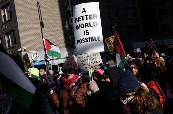 A crowd participating in a protest, holding various signs. Notable among the signs is one that states, "A BETTER WORLD IS POSSIBLE." Flags, including those representing Palestine, are visible, and participants are dressed warmly for the cold weather.