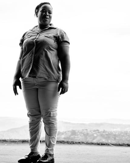 A person stands confidently in front of an expansive landscape, wearing a button-up shirt and pants. The image is in black and white, highlighting the contrast between the person and the bright background of distant hills.