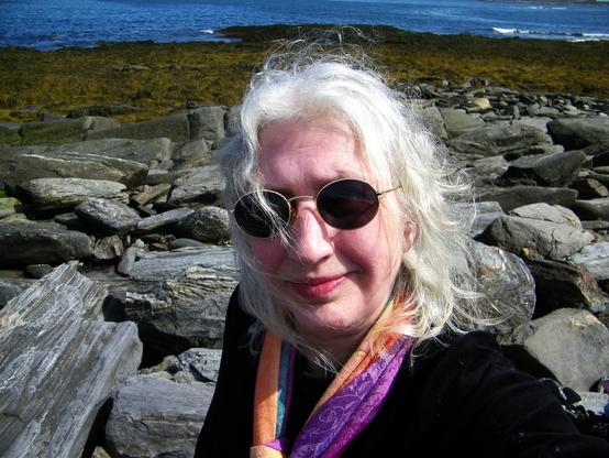 A person with white hair and pale skin, wearing dark sunglasses and black clothing, but a colorful pastel rainbow scarf. In the background are rocks, seaweed, and the ocean -- blue with some white waves.