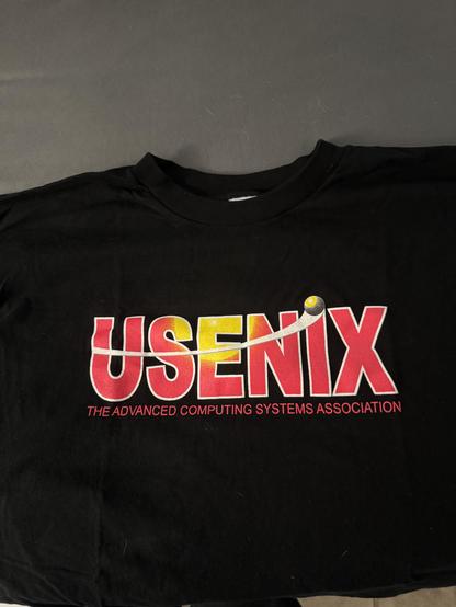 Black T-shirt on a gray background. The shirt features a logo in red/orange and yellow that says USENIX in all caps, above the legend (smaller, also all caps) in red reading THE ADVANCED COMPUTING SYSTEMS ASSOCIATION.