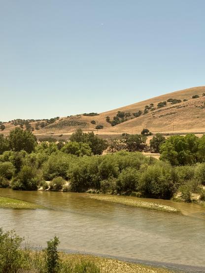very shallow river running alongside the train with dark green trees on the far bank and yellow hills behind it