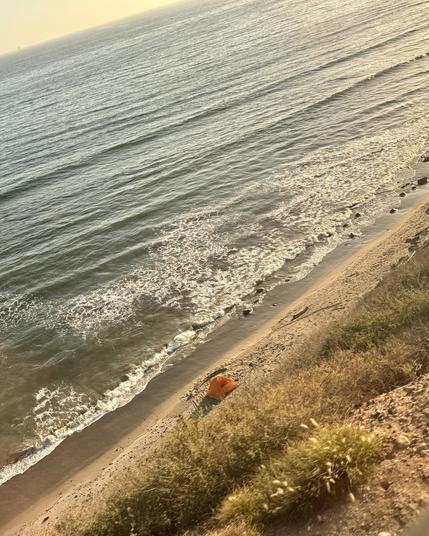 view from the top of a cliff down to the beach. The ocean is a gray blue and the beach is narrow. There’s a bright orange tent set up on the beach.