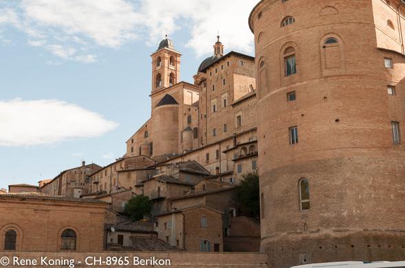 Das Bild zeigt eine historische Stadt (Urbino) in Hanglage mit einem markanten architektonischen Stil. Die Gebäude sind aus warm getönten Ziegeln errichtet und verleihen der Stadt ein einheitliches und traditionelles Aussehen. Im Vordergrund ist ein großer zylindrischer Turm mit Rundbogenfenstern und einer Kuppel zu sehen, der auf ein befestigtes oder bedeutendes Gebäude hindeutet. Dahinter erhebt sich eine Reihe von Gebäuden auf dem Hügel, aus denen eine Kirche mit Glockenturm und Kuppel hervorsticht. Der Glockenturm der Kirche hat bogenförmige Öffnungen, und die Kuppel ist mit einem Kreuz versehen. Die Gebäude stehen dicht beieinander, mit Terrakotta-Dächern und kleinen Fenstern, typisch für die mittelalterliche europäische Architektur. Der Himmel ist teilweise bewölkt, und das Sonnenlicht wirft Schatten auf die Gebäude und hebt ihre Strukturen und Details hervor. Die gesamte Szene vermittelt ein Gefühl von historischer Tiefe und architektonischer Harmonie.
*** Übersetzt mit www.DeepL.com/Translator (kostenlose Version) ***