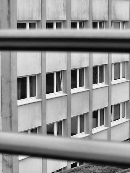 Schwarzweißfoto durch zwei horizontale Lamellen einer Jalousie am Zimmerfenster auf einen schräg gegenüberliegenden mehrstöckigen Plattenbau mit vielen Fenstern.
