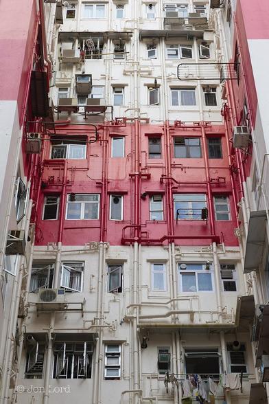 This Is A Colour Cityscape / Abstract Photo In Landscape Format. Rising From The Base To The Top Of The Frame Is The Back (Front Or Side ?) Of An Older Apartment Building With Seven Floors In View. The Main Colour Is Cream But With A Bright Red Stripe Running Horizontally Across Two Floors Midway Up The Building. There Is Some Symmetry With The Multitude Of External Pipes Mostly Being Aligned Vertically Or Horizontally But There The Order Stops. Some Of The Aluminium Framed Windows Are Square, Some Rectangular, Some With Criss Crossed Bars For Security, Some Without. Some Have External Air Conditioning Units, Some Have None - Some Have Two. Some Have Clothing Drying, Some Not. 
Concluding: A Very Typical (But Recently Painted) Older Low-Rise Hong Kong Apartment Block, This One Located In Causeway Bay (2013).