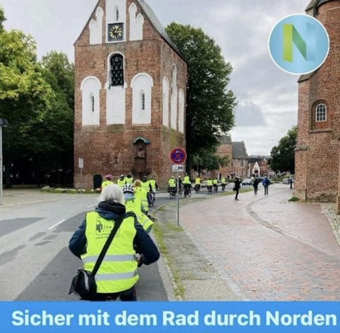 Viele Fahrradfahrende mit gelben Warnwesten fahren durch Norden vorbei am Glockenturm der Norder Ludgerikirche. Im Hintergrund sieht man das Ostfriesische Teemuseum, die Westerstrasse und die alten Bäume des historischen Marktplatzes