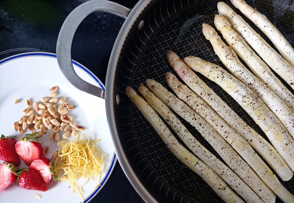 rechts eine Pfanne mit weißem Spargel, links ein Teller mit gerösteten Pinienkernen, Zitronenzesten und halben Erdbeeren
