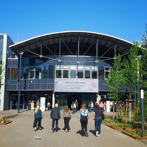 View of a modern university building under a clear blue sky, with people walking toward the entrance. A large banner above the entrance reads "Herzlich willkommen zur Festveranstaltung der H-BRS," indicating a festive event at Hochschule Bonn-Rhein-Sieg.