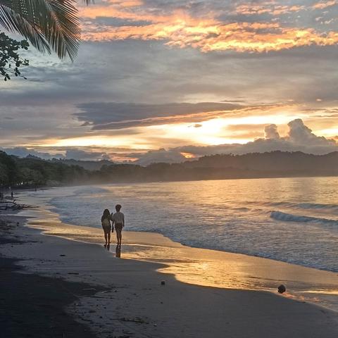 Two young black women walk on a golden wet beach, reflecting the sunset in the background.