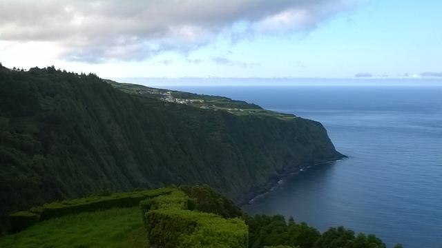 The image depicts a coastal landscape with a steep, lush green cliffside descending to the calm blue ocean. In the foreground, there is a neatly trimmed hedge, adding a structured element to the natural scene. The cliff is densely covered with greenery, indicating a rich, vegetated area. The ocean is visible in the background, with its surface reflecting the sky, and the horizon is visible where the sea meets the sky. The sky is partly cloudy, with patches of blue visible through the clouds. In the distance, a small settlement or village can be seen atop the cliff, with buildings that appear to be white or light-colored, blending into the landscape. The overall scene is serene and picturesque, showcasing the natural beauty of the coastal region.