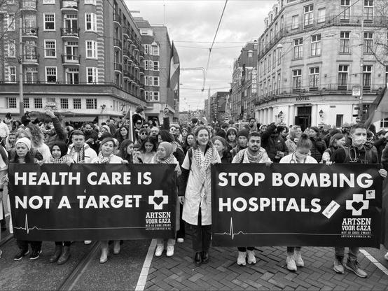 A black and white photograph of a protest march in Rotterdam, Netherlands, against Israel's actions. A large group of nurses is walking down a city street, holding two prominent banners. 

The banner on the left reads "HEALTH CARE IS NOT A TARGET" and the one on the right says "STOP BOMBING HOSPITALS." 

Both banners have the logo and text "ARTSEN VOOR GAZA" (Doctors for Gaza)with a heart symbol and a heartbeat line graphic. 

The crowd consists of people of various ages and backgrounds, some wearing scarves and winter clothing. The street is lined with tall buildings, and the atmosphere appears peaceful yet determined.