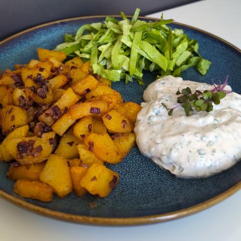 Picture of a meal on a blue plate with three components. It consists of fried potatoes and red onions seasoned with smoked pepper, a vegan almond curd with herbs garnished with a small bunch of sprouts and a portion of plain green lettuce. The background is a neutral white table.