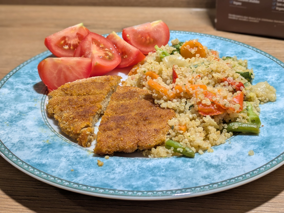 Das Bild zeigt einen bunten, veganen Teller mit drei Komponenten:

- Vorne links: Ein paniertes, veganes Schnitzel, das in zwei Hälften geschnitten wurde. Die goldbraune, knusprige Panade und die lockere Struktur deuten auf eine selbstgemachte oder hochwertige pflanzliche Variante hin.
- Rechts: Eine Portion Quinoa-Gemüse-Pfanne, bestehend aus gegarter Quinoa und einer bunten Mischung von Gemüse – zu erkennen sind Karotten, grüne Bohnen, Paprika, Zucchini und mehr. Die Farben sind kräftig, das Gericht wirkt leicht und sättigend.
- Oben links: Einige Stücke frischer Tomate, geviertelt, saftig und roh serviert – eine frische und einfache Ergänzung.

Der Teller hat, wie viele deiner Gerichte, einen grünen Rand mit Muster und steht auf einer hellen Holzarbeitsfläche. Die Mahlzeit ist gesund, ausgewogen und ansprechend angerichtet.