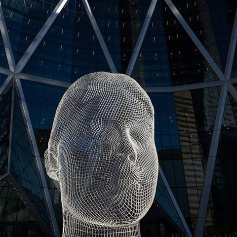 Photograph of a white wireframe sculpture of a human head, placed in front of the triangular glass frames of a large residential building.