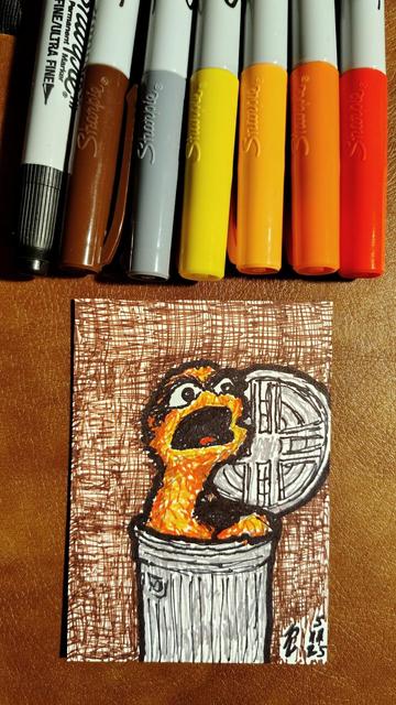 Marker drawing of the original 1969 version of Oscar the Grouch (colored yellowish-orange with black eyebrows instead of his later green with brown eyebrows) hanging out in his trash can. The drawing is signed with my "RV" signature glyph and dated 5-11-25. The black, brown, grey, yellow, light orange, dark orange, and red markers used to draw the image rest on the table above the card.