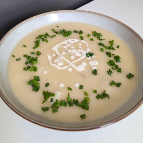 Picture of a vegan asparagus cream soup with small barely visible pieces of asparagus on an gray plate. The soup is of light beige color and seasoned with chives and a splash of oat cream. The background is a neutral white table.