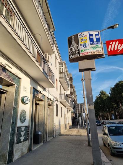 A street scene featuring a "Bar Cafe de Paris" sign, various advertisements on a large sign, and a parked car. The buildings are light-colored with balconies.