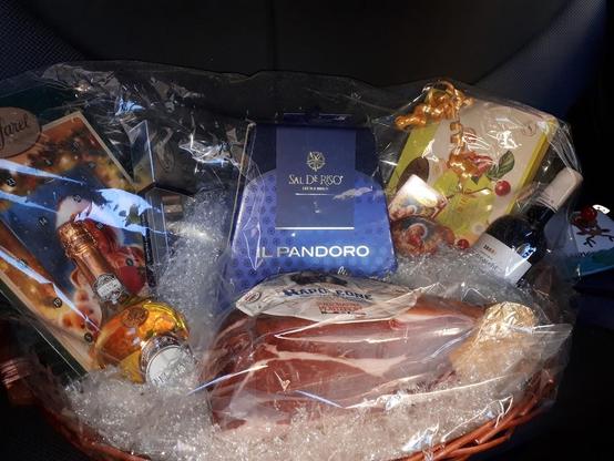 A gift basket is filled with items like ham, a bottle of wine, a blue box with the words "Sal de Riso," a small bottle of sparkling wine, a box with a Christmas advent calendar, and other wrapped gifts, all covered in clear plastic wrap and resting in a wicker basket.