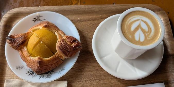 A wooden tray with a pastry and a coffee. The pastry is a square with a yellow fruit in the middle. The coffee is in a white geometric cup.