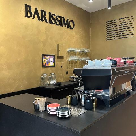 A brightly lit cafe interior with a "BARISSIMO" sign on a gold wall. A black countertop holds coffee-making equipment, colorful plates, and utensils. The menu board lists various coffee drinks and prices.