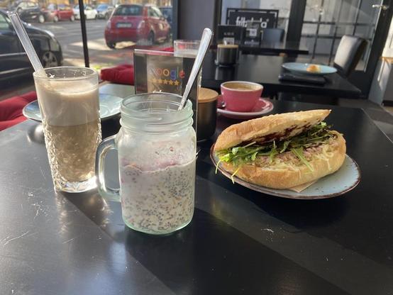 The image shows a table with a latte, a chia seed jar, a sandwich, and a coffee cup.