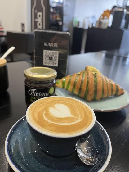 A coffee with latte art, a green pistachio croissant, and a jar of "Oliviata" jam on a black table.