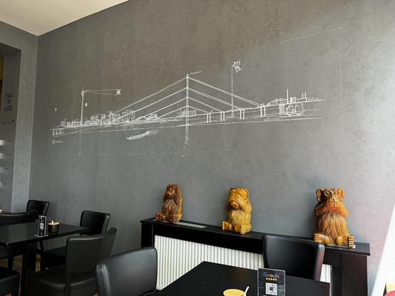 A gray wall with a white drawing of a bridge and cityscape. Three wooden monkey statues sit on a black cabinet in front of the wall. Tables and chairs are visible in the foreground.