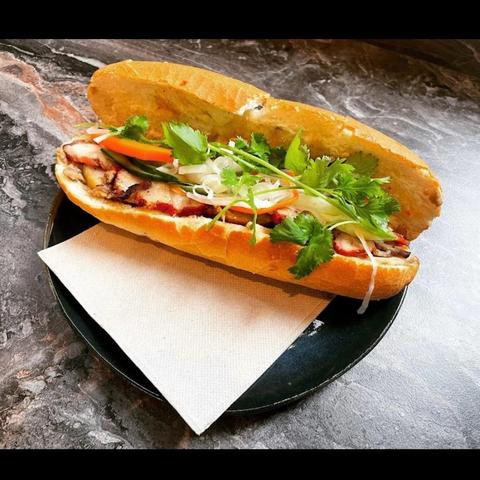 A banh mi sandwich sits on a plate with a napkin, featuring grilled pork, pickled vegetables, and herbs.