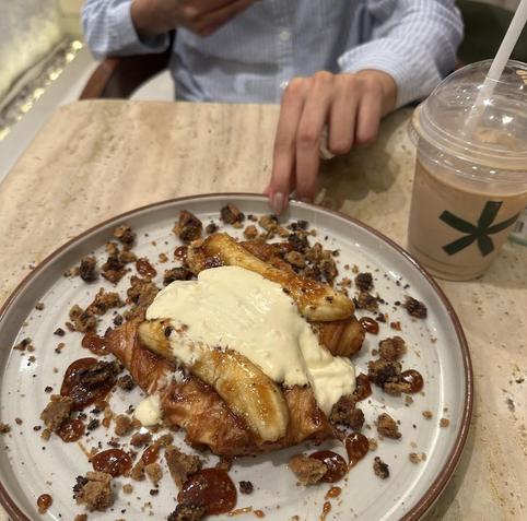 A croissant topped with caramelized banana slices and a scoop of cream is surrounded by chocolate crumbles and caramel sauce on a white plate next to a beverage.
