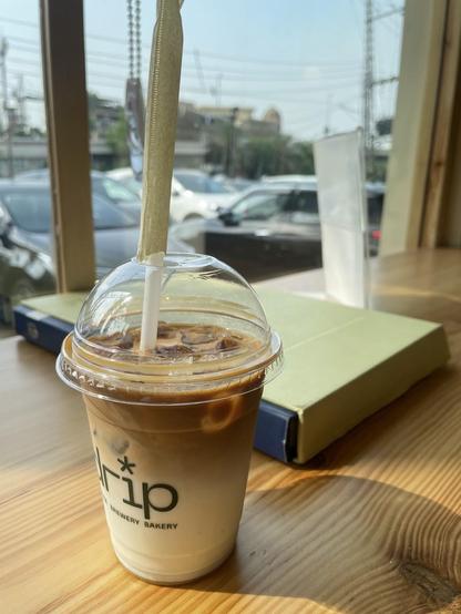 Iced coffee with straw on a wooden table, books behind. "Drip Brewery Bakery" logo on the cup. Natural light.