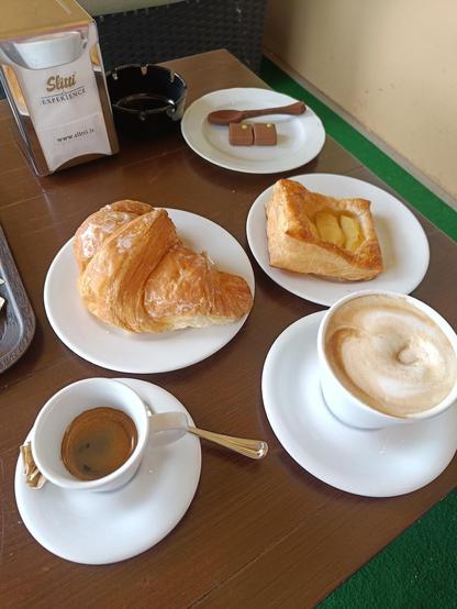 A table with a croissant, a pastry, espresso, cappuccino, chocolate, wooden spoon, and an ashtray.