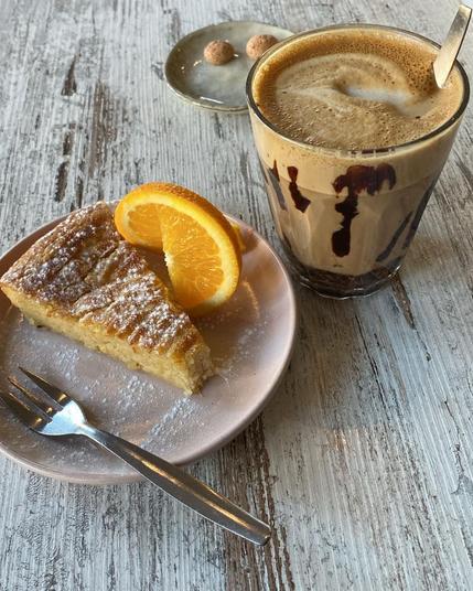 A slice of cake with an orange slice, a glass of coffee, and a fork on a wooden table.
