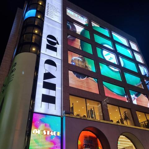 Here's a description of the image:

A multi-story building featuring a large digital display showing a record player with a green vinyl record. A sign reading "OI CAFE" is attached vertically to the building. Another LED display shows the words "OI STAGE" in colorful lettering.