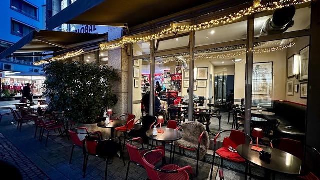 A charming outdoor cafe with red chairs and tables, lit by warm lights.