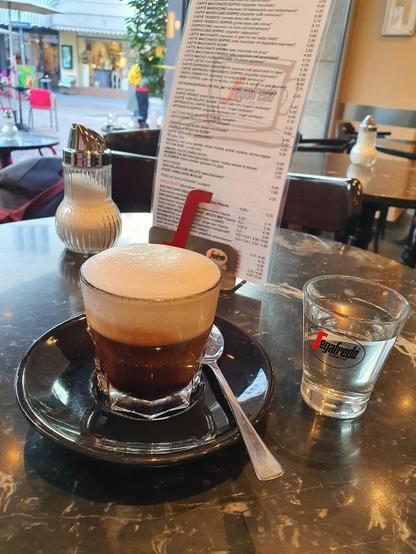 A cup of coffee with foam on a black saucer, accompanied by a small glass of water, sits on a marbled table in front of a menu.